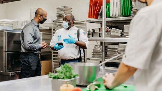 African American Service Rep meeting with restaurant manager in kitchen using Ecolab Sales & Service on phone.